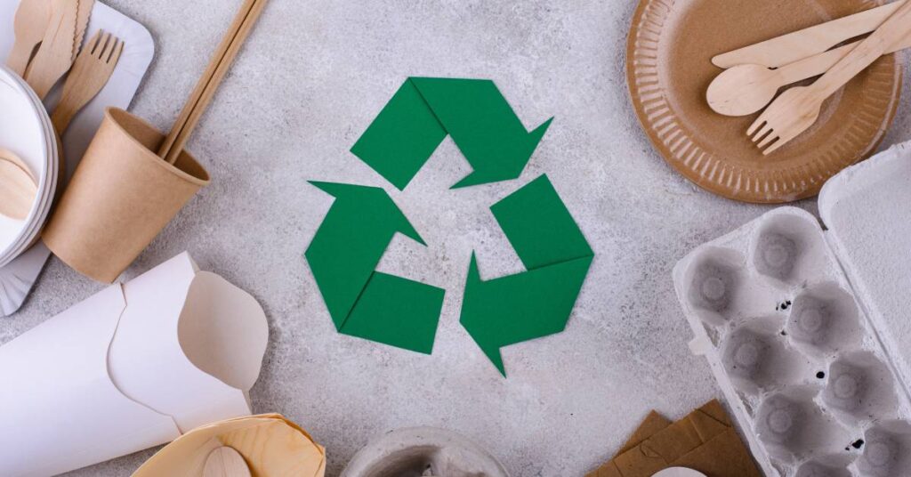 A green recycling logo surrounded by various recyclable products such as egg cartons, paper cups, and wooden dishware.