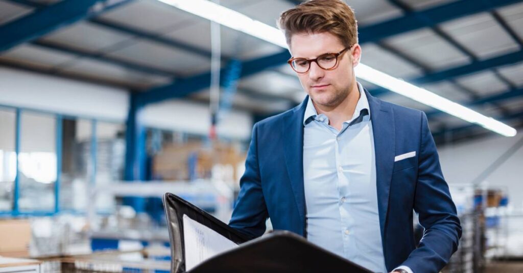 A businessman wearing a blue suit and glasses standing in a warehouse reading a portfolio beneath a light.