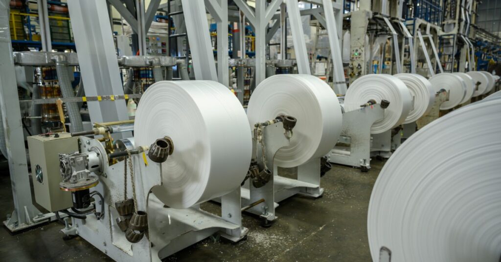 A row of machines on a factory floor with plastic film fed into them. There’s a scaffolding in the background.