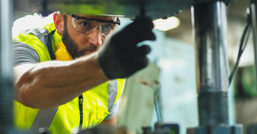 A maintenance worker in a factory wearing a high-vis vest and goggles operating on a piece of equipment.