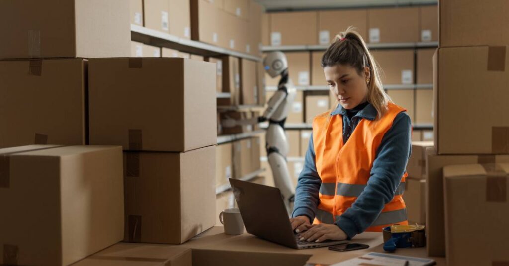 A woman wearing an orange vest working on a computer in a room full of boxes, with a robotic worker in the background.