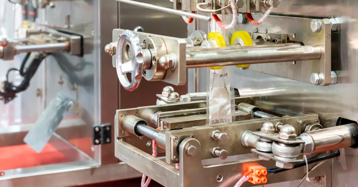 A large industrial machine with various tubes and gears filling a pouch with white powder on a manufacturing floor.