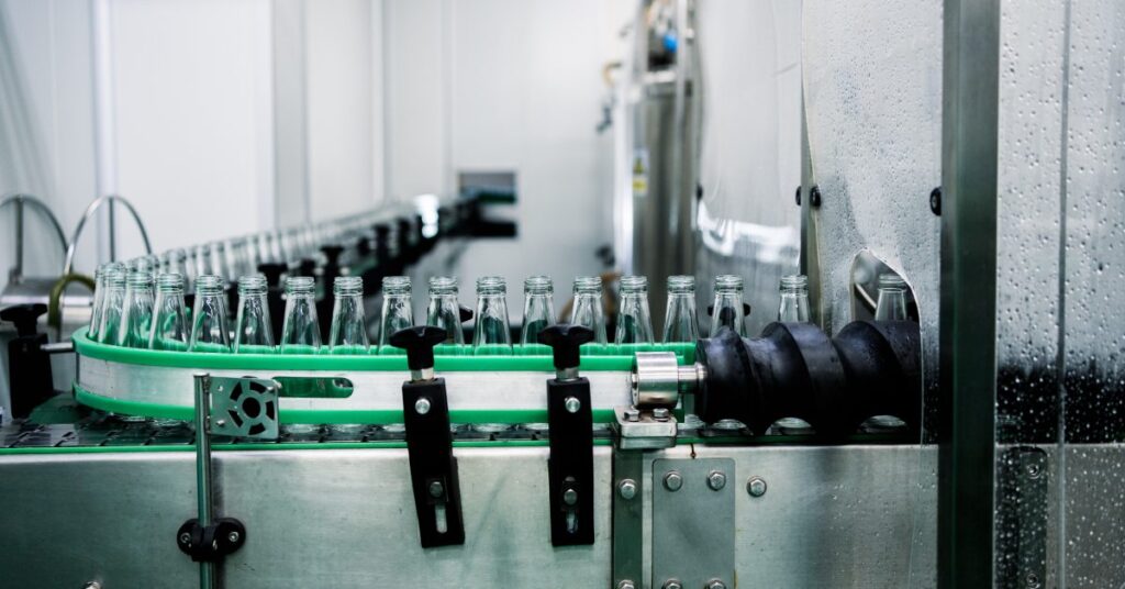 A line of glass bottles moving along a conveyor belt through a rotary filling machine on a manufacturing floor.