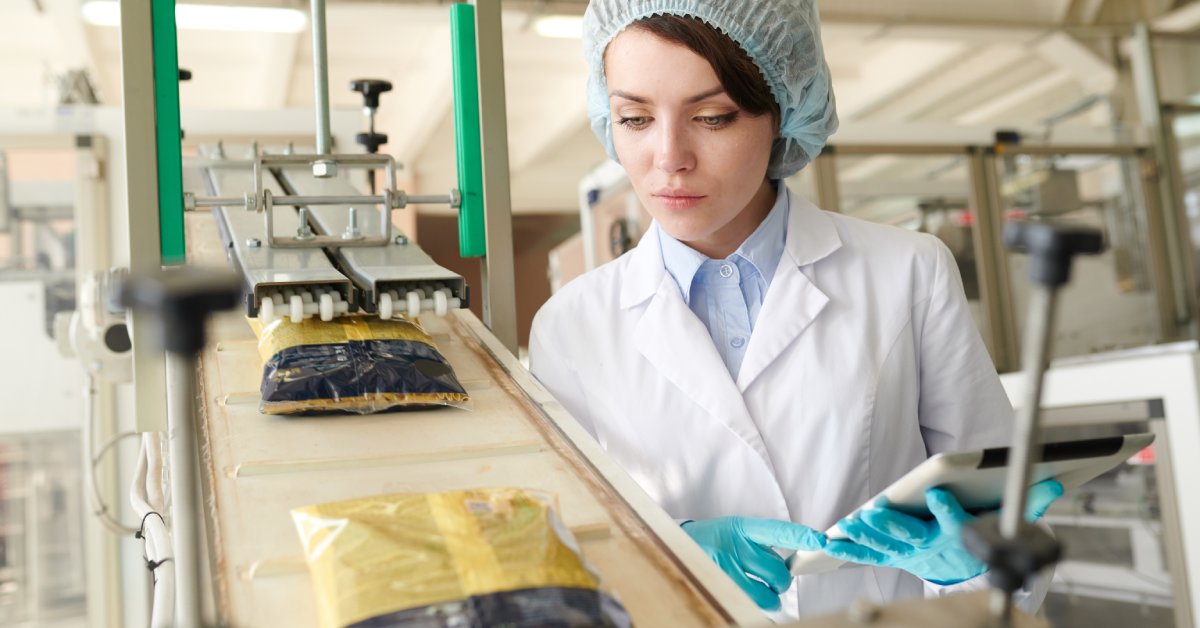 A woman in a white coat and hairnet holding a clipboard while inspecting food packaging coming out of a fill machine.