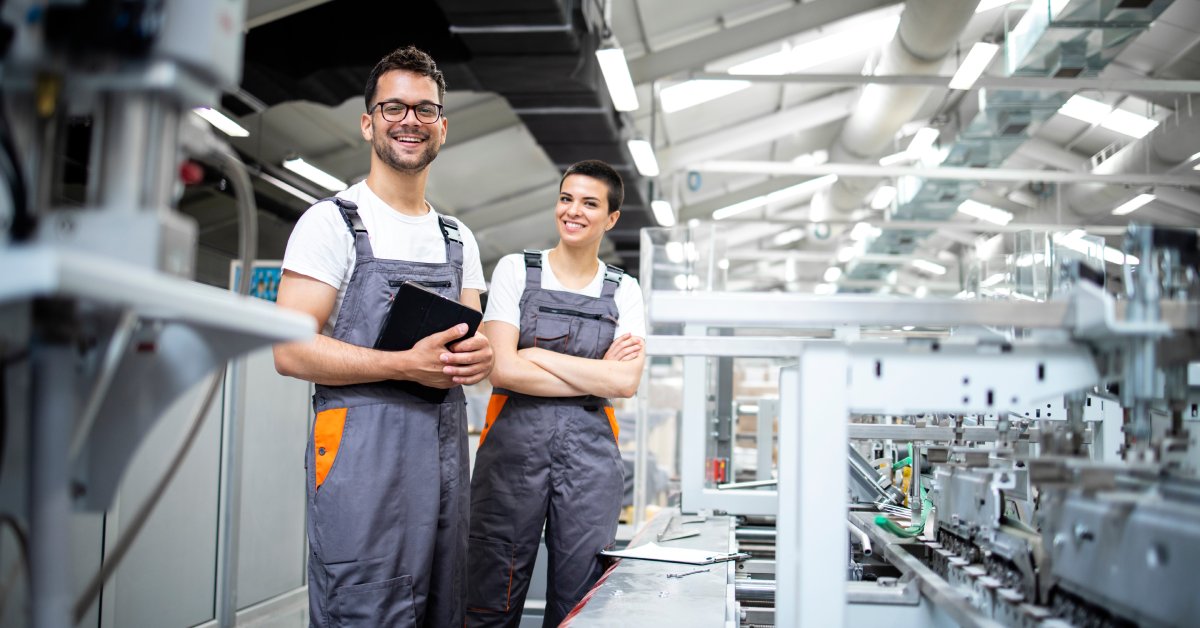 Two workers wearing gray overalls and white t-shirts smiling while standing over a production line in a factory.