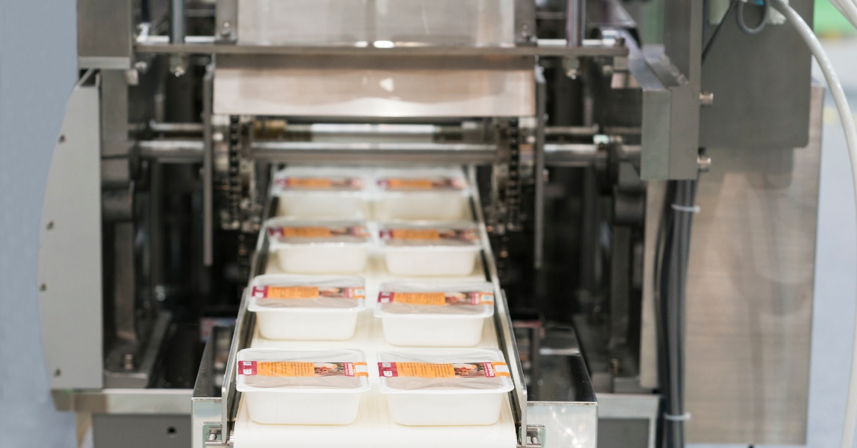 A conveyor belt with packaged food product trays coming out of an industrial packing machine side-by-side.