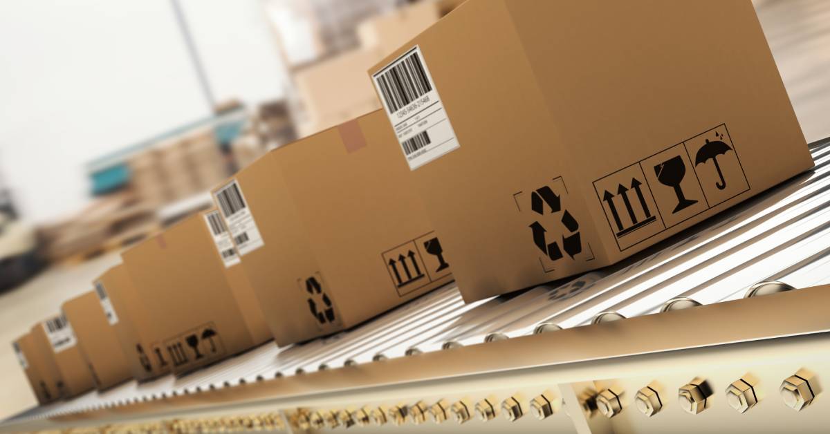 A row of boxes with barcodes and black recycling symbols on them moving down a conveyor belt on a factory floor.