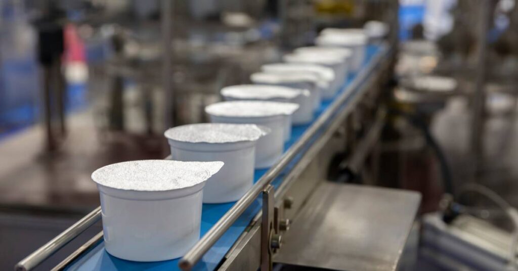 Individual white cups with sealed foil lids moving down a blue conveyor belt on a packaging line in a warehouse.