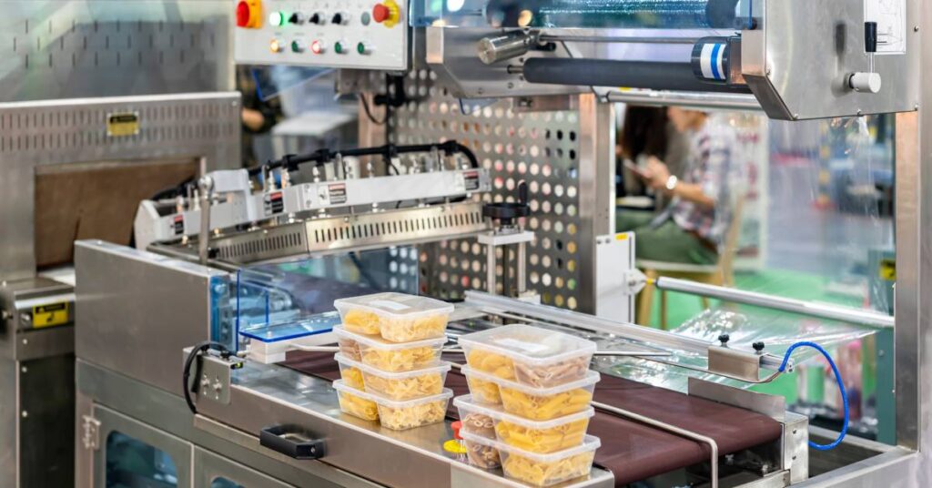 Food in plastic boxes sitting on an automatic machine with a conveyor belt. There is a person in the background sitting.