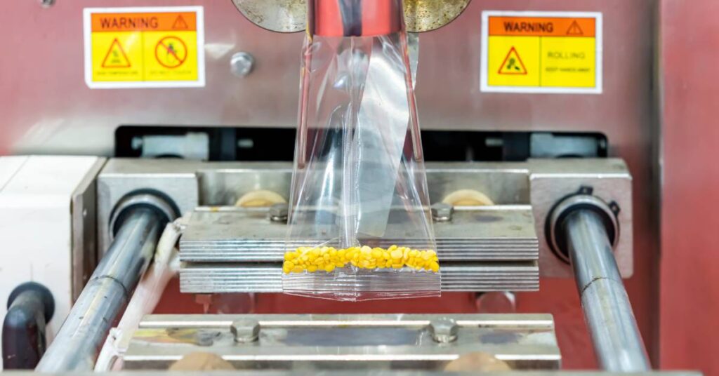 A large industrial packaging machine fills a plastic bag with yellow granules. The machine has two warning stickers.