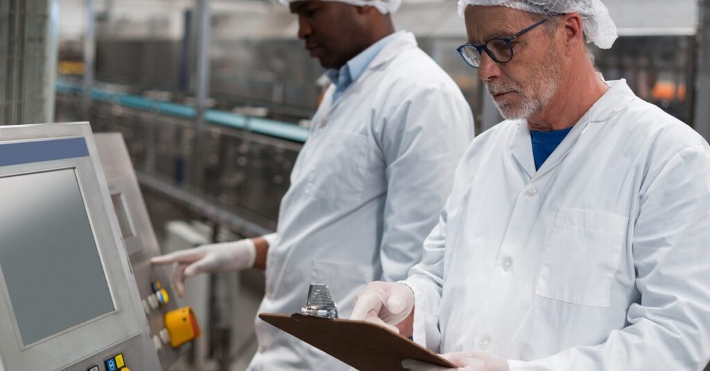 Two factory workers wearing hairnets inspect equipment. One takes notes on a clipboard. There is a conveyor belt behind them.