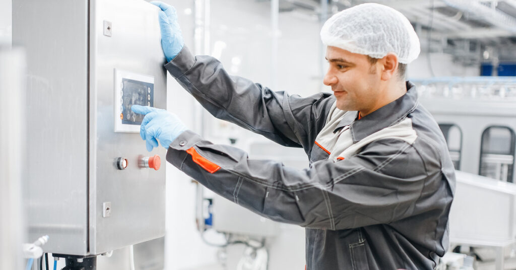 A man wearing a hairnet operates the controls of a machine in a brightly lit and clean production plant.