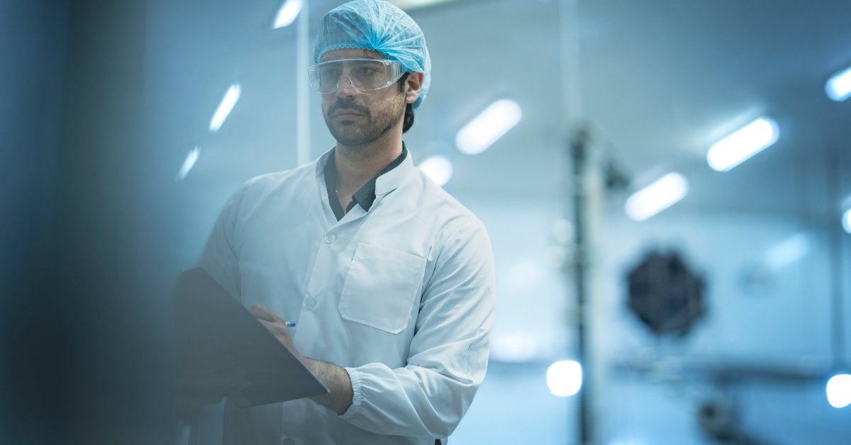 A man wearing a hairnet and protective glasses takes notes on a clipboard. The background is blurred.