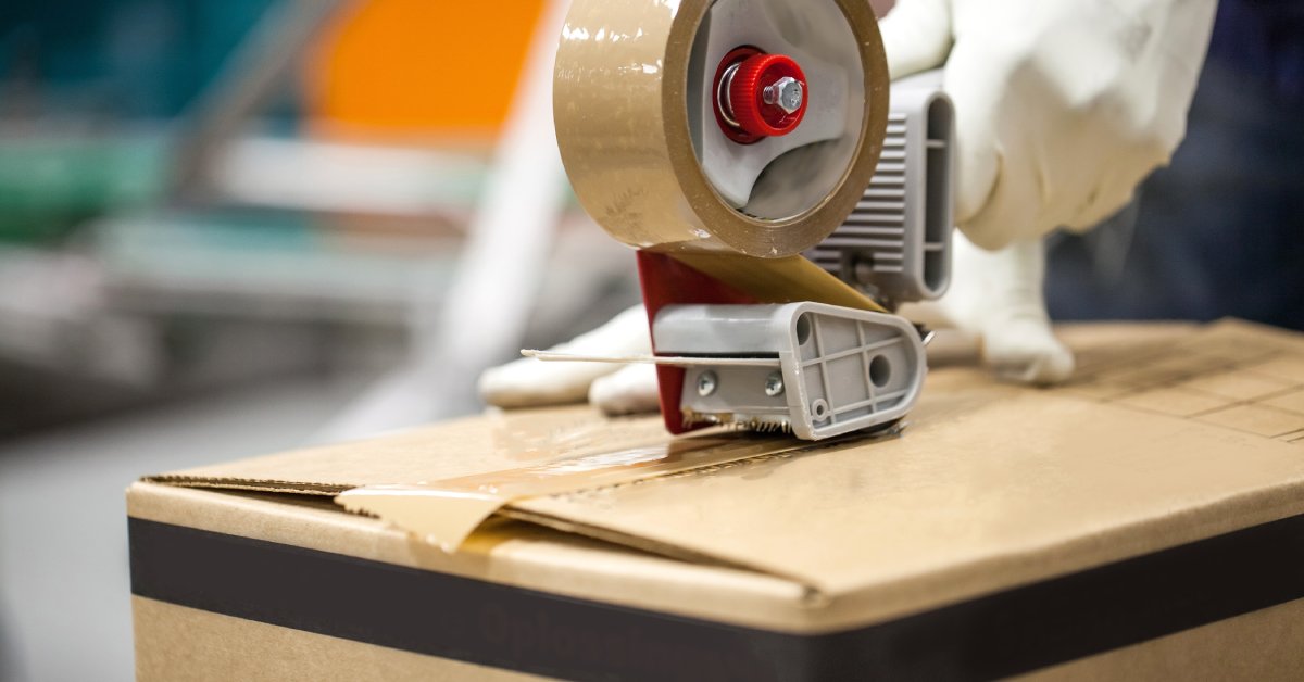 A person wearing gloves and using a tape roller to seal the top of a cardboard box on a packaging plant floor.