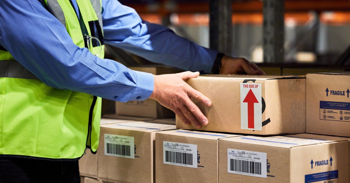 A warehouse worker organizes boxes for shipment. Products are labeled with barcodes and safety instructions.