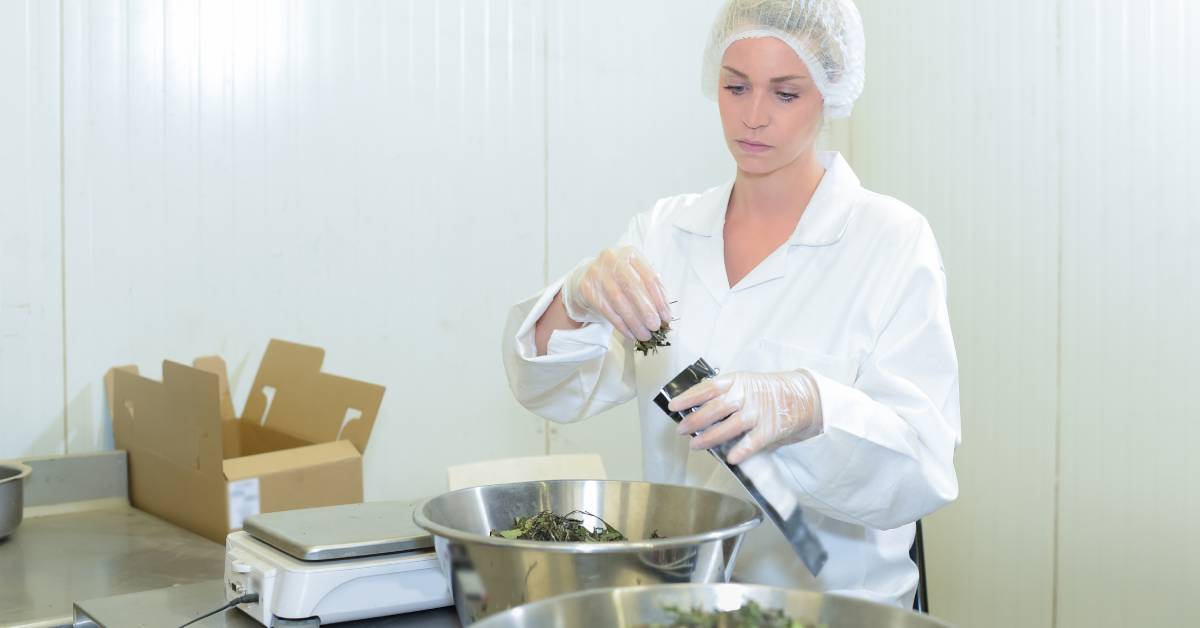 A woman wearing a hairnet and gloves stands at a table with a scale and fills a silver pouch with a product by hand.